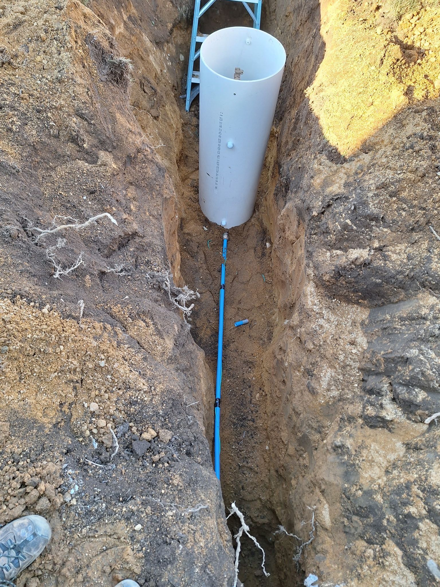 Trench with a tall white pipe and blue piping running along the bottom. Brown earth surrounds.