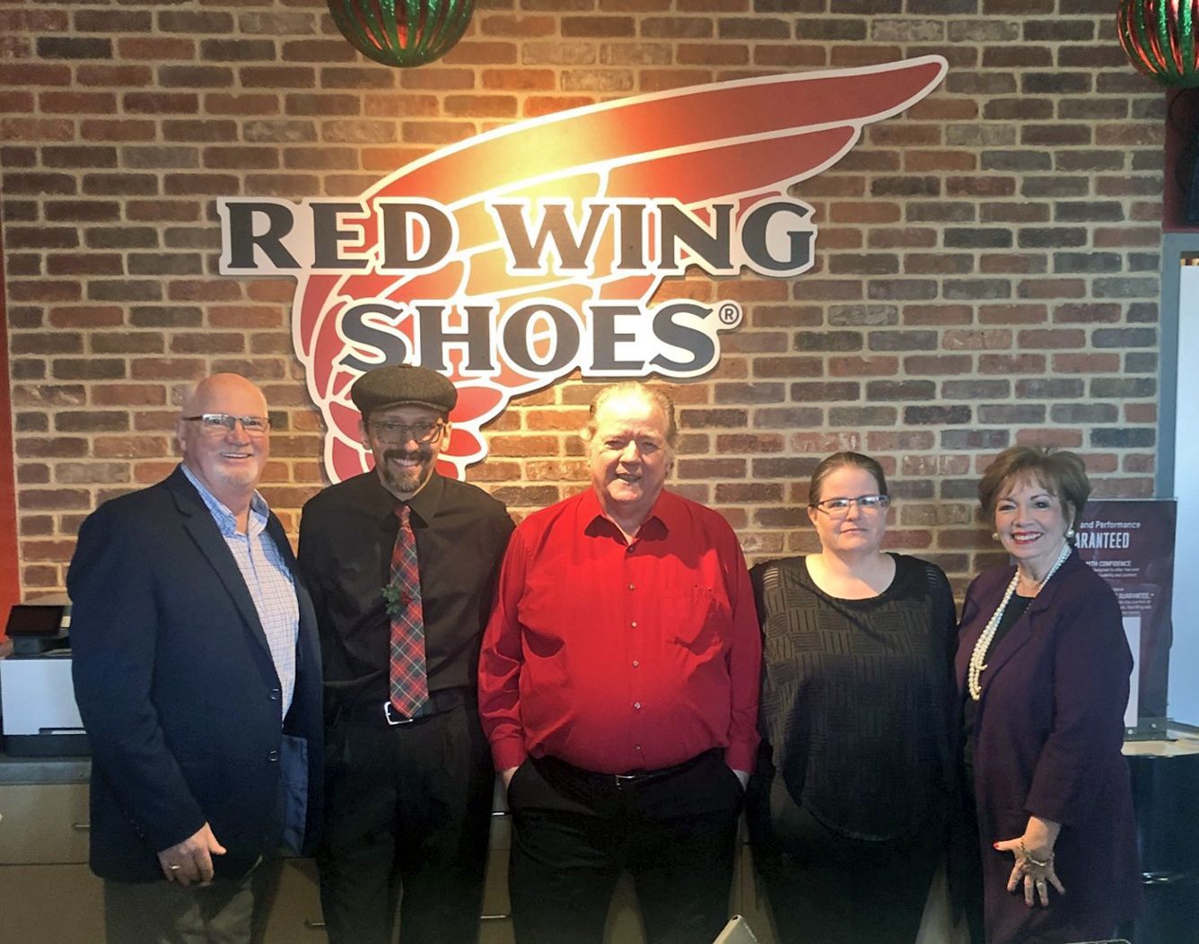 Five people smiling in front of a Red Wing Shoes logo on a brick wall.