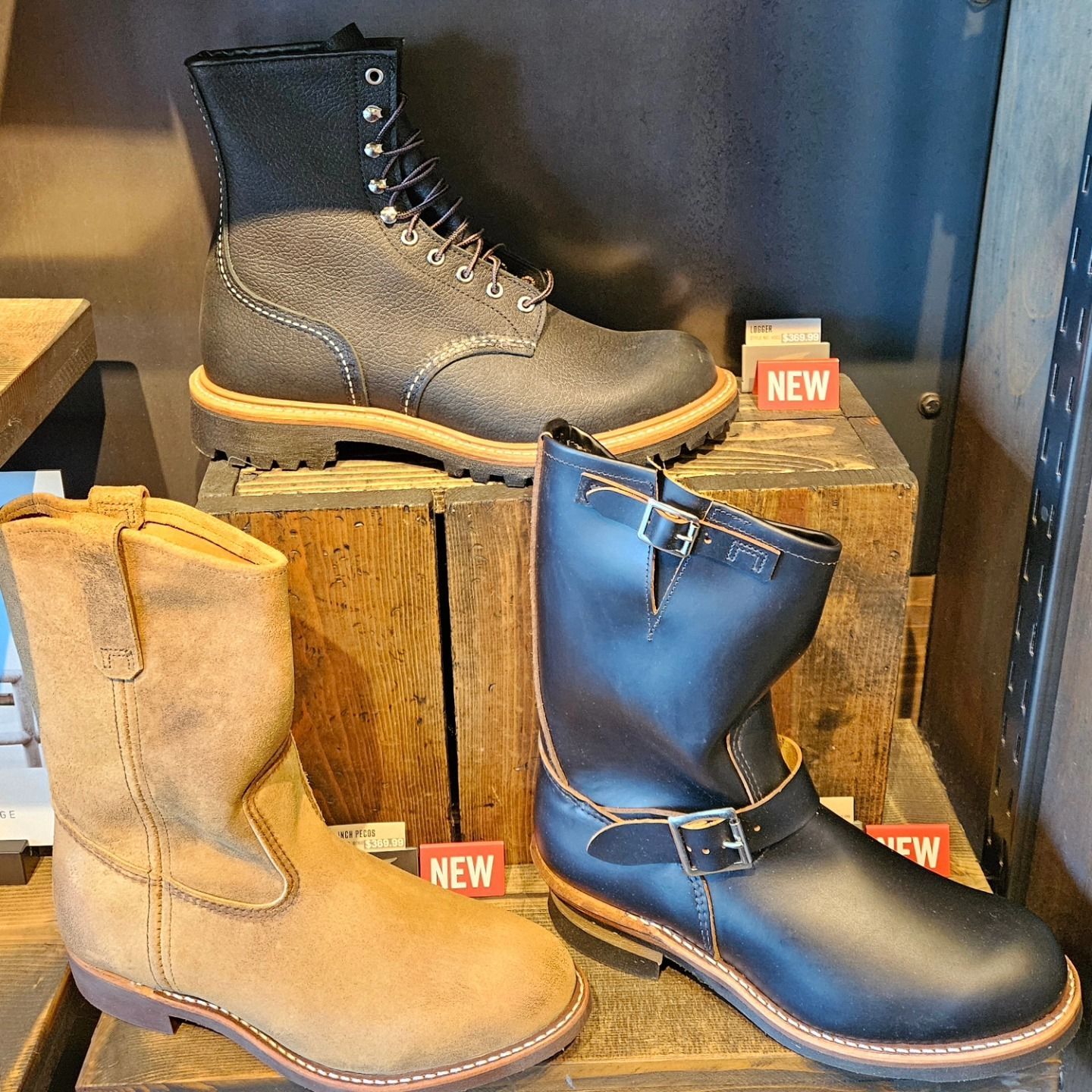 Three leather boots displayed on wooden crates: brown, black, and gray with laces and buckles.
