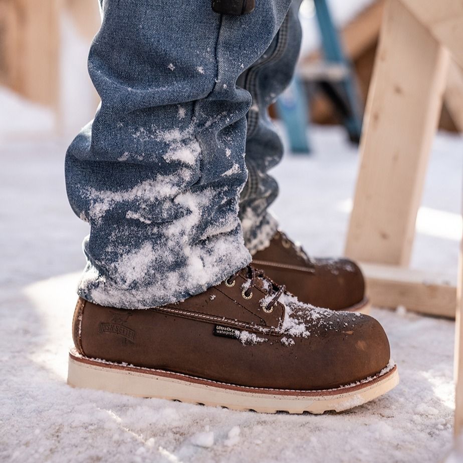 Person wearing work boots and jeans covered in snow outdoors near wooden structures.