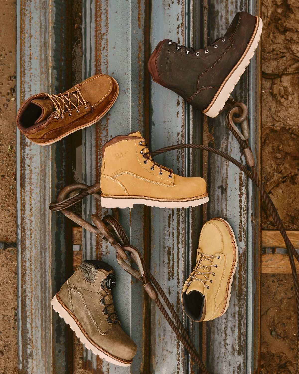 Collection of work boots in various colors (browns, tan, and olive green) arranged on a weathered metal surface.
