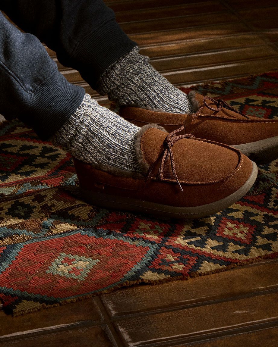 Feet in brown suede slippers and gray knit socks on a patterned rug.