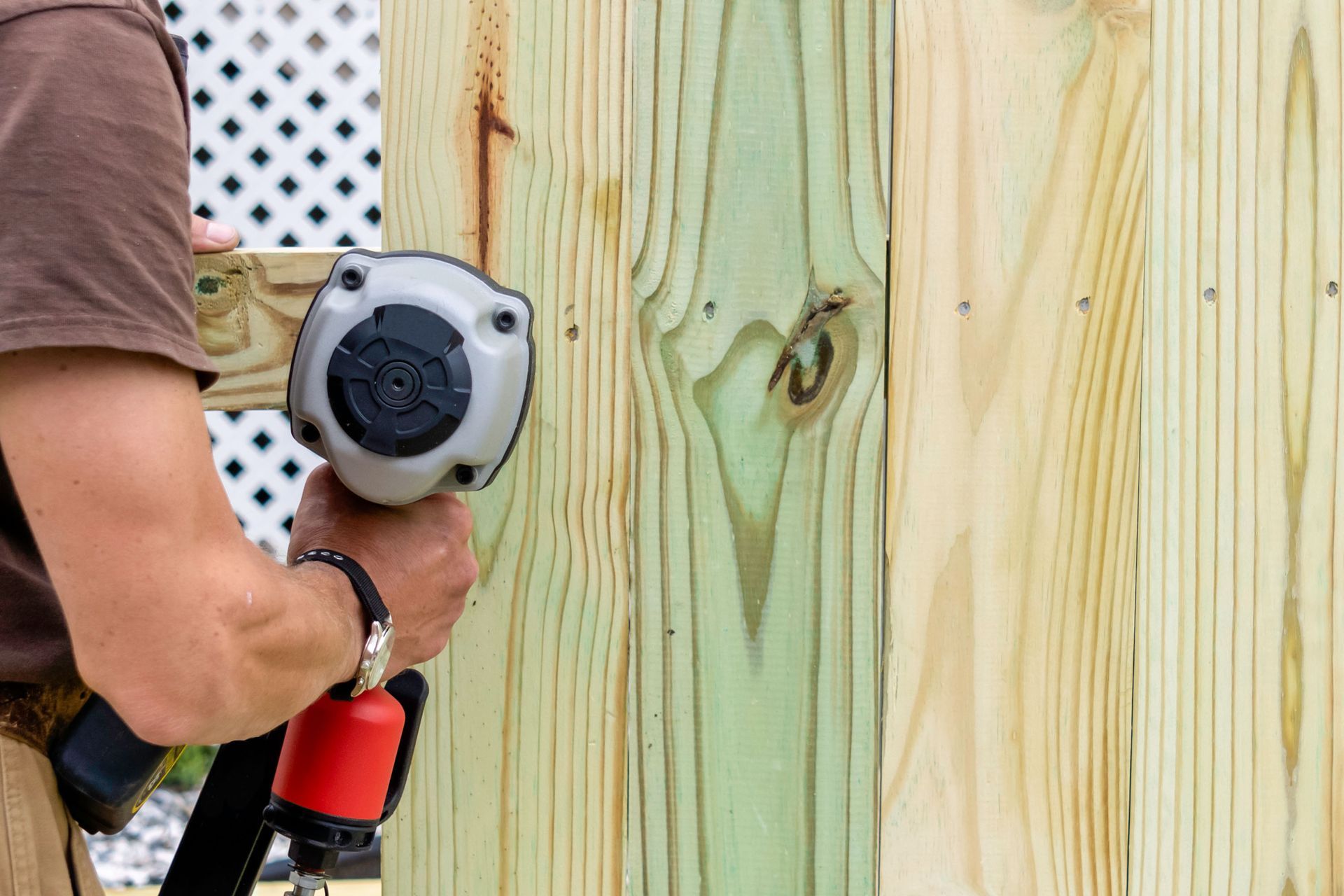 Person using a nail gun to fasten wood planks together, building a fence outdoors.
