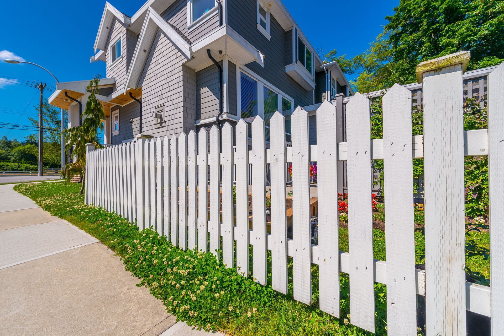 White picket fence in front of a gray house with a sidewalk and grass.