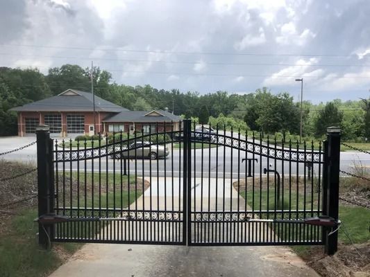 Black metal security gate on a concrete driveway, leading to a brick building on a cloudy day.