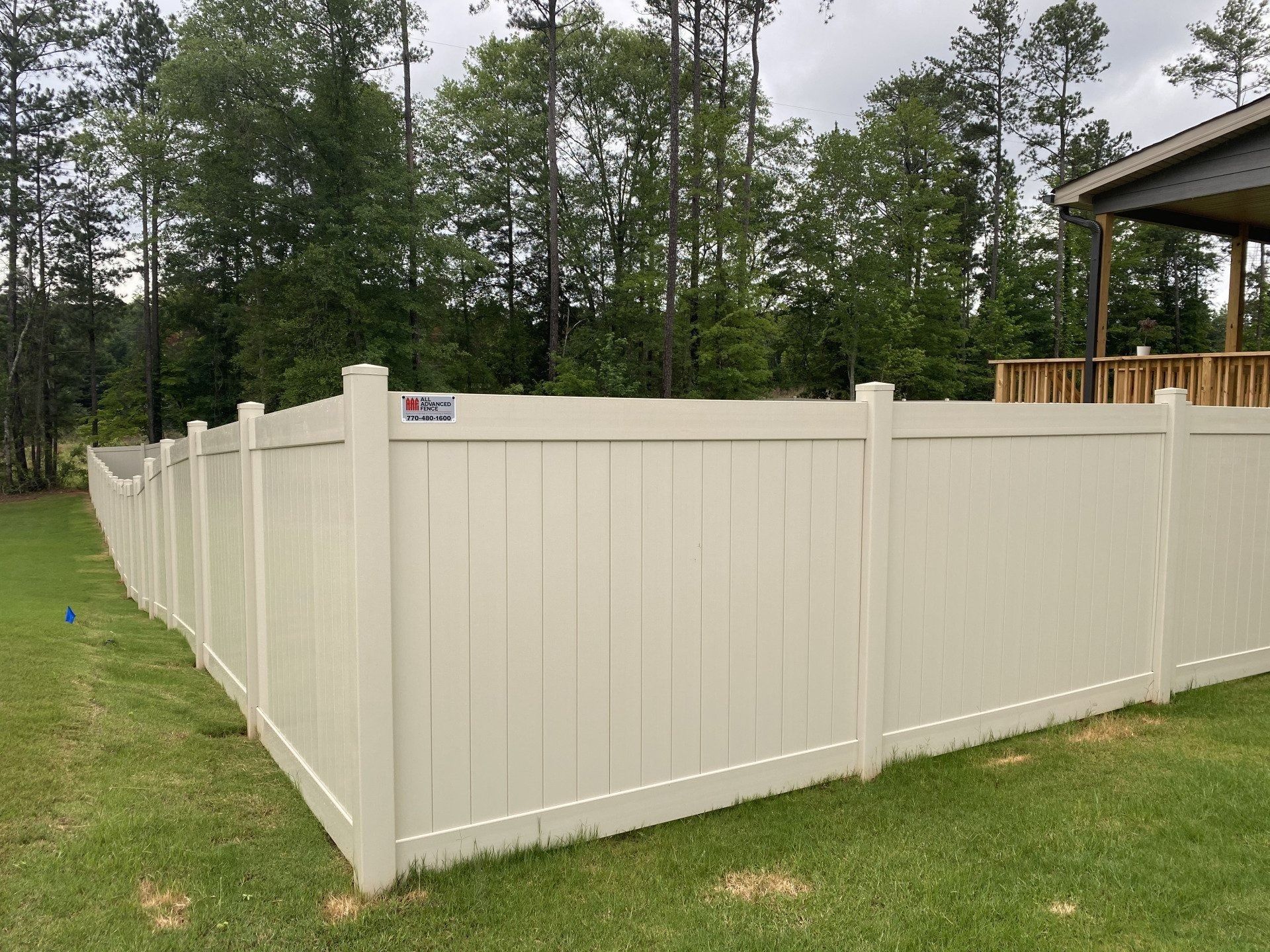 A tan vinyl privacy fence surrounds a green lawn, with a wooded background and part of a house.