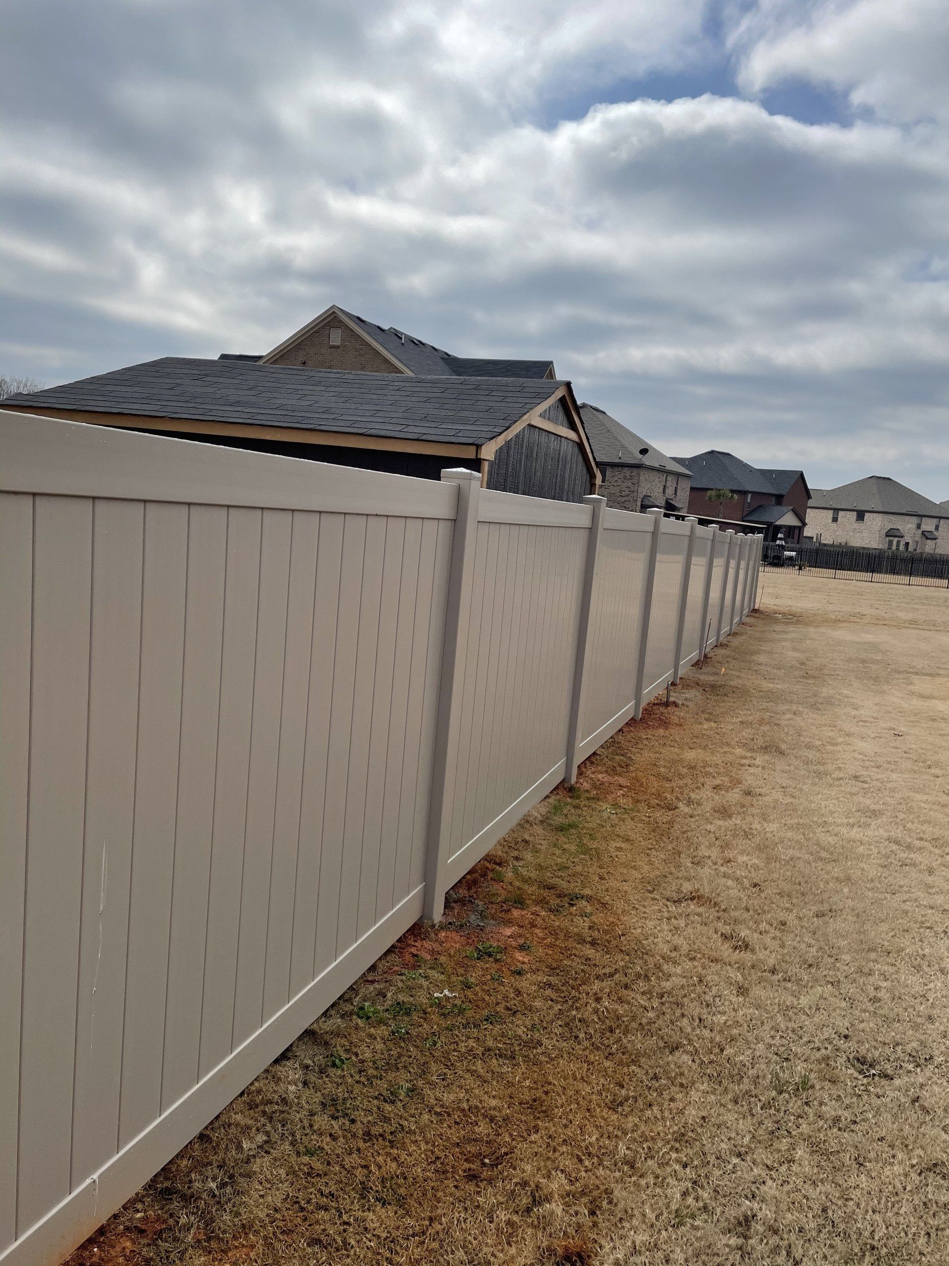 Tan vinyl fence in a residential area, with houses in the background under a cloudy sky.