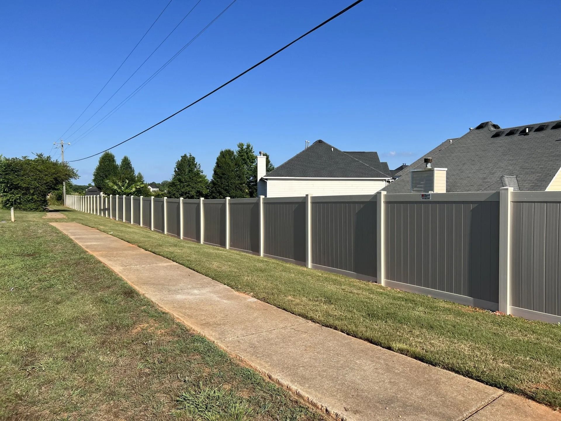 A long gray fence bordering a sidewalk and grassy yard, with houses in the background under a blue sky.