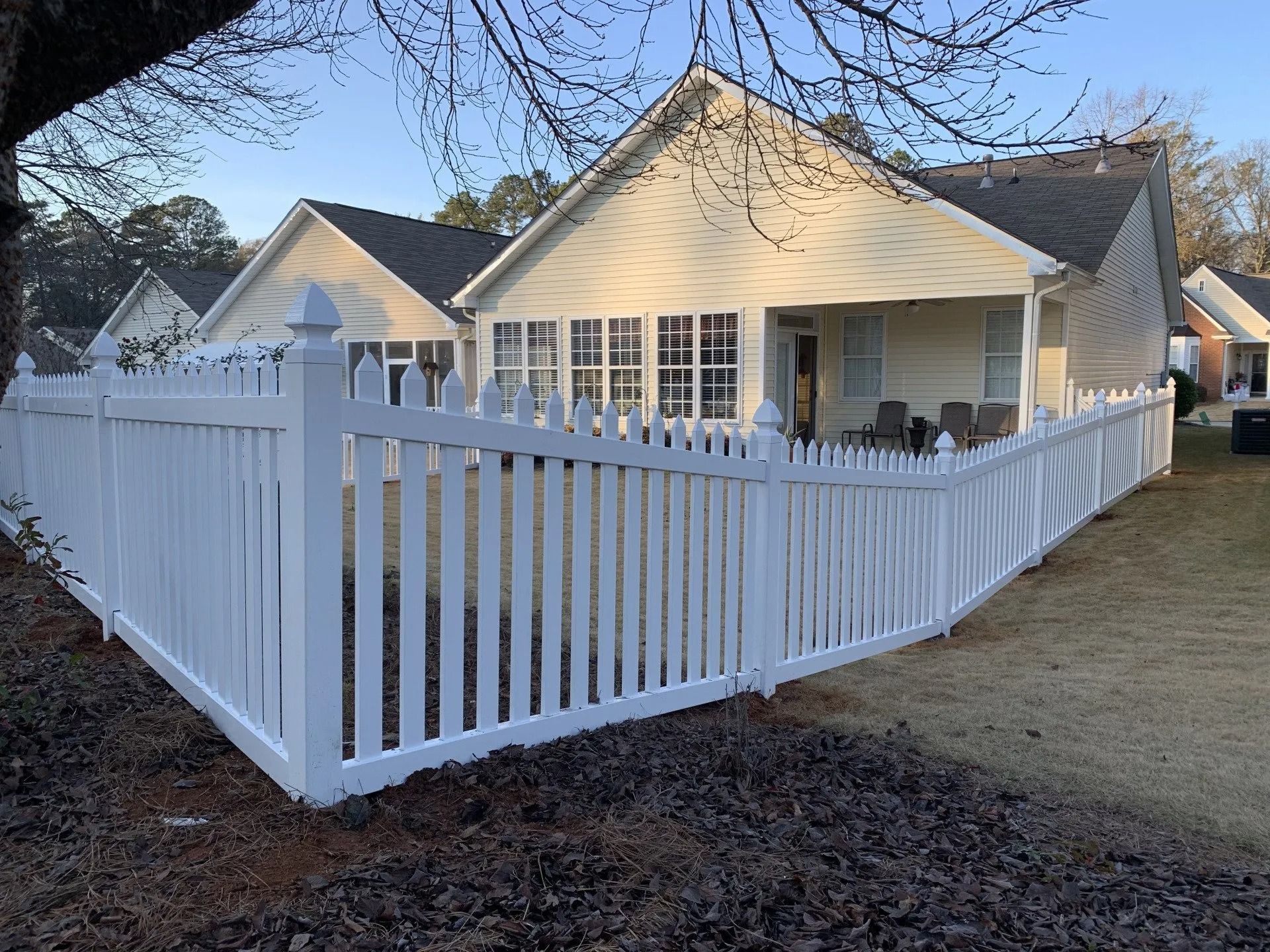 White picket fence surrounds a light-colored house with a dark roof.