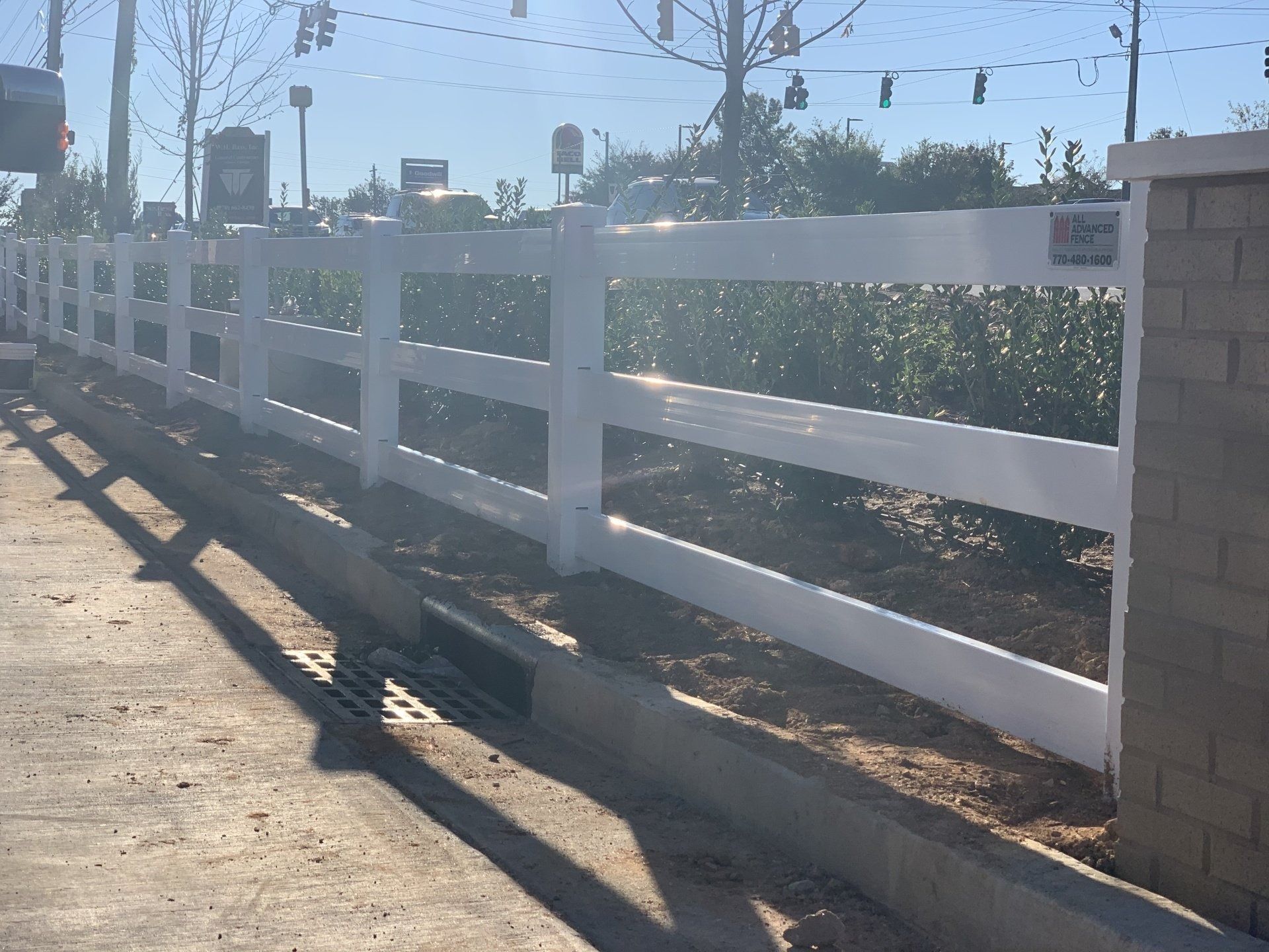 White, three-rail fence alongside a concrete curb, near a street with traffic signals and a building.