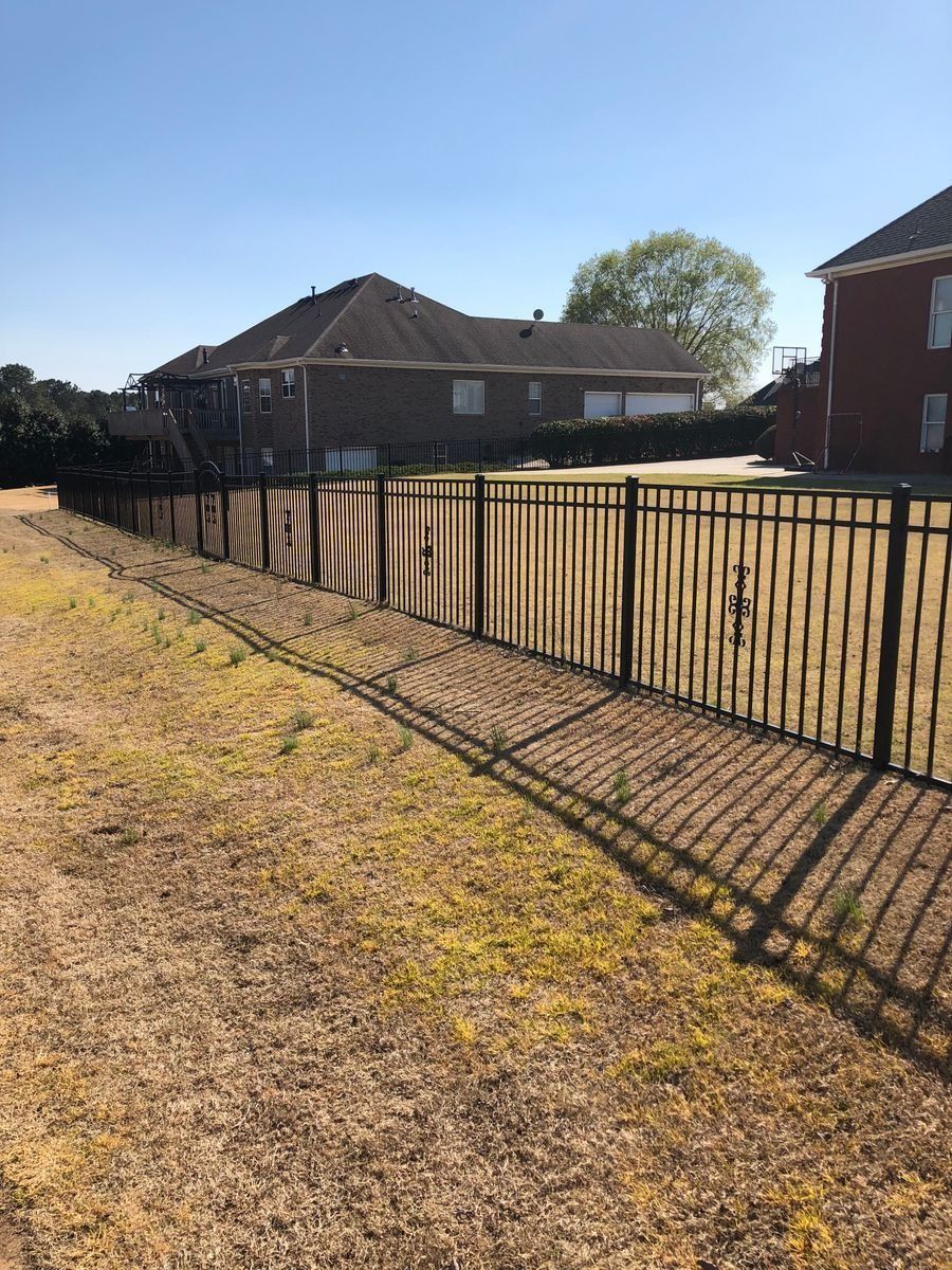 Black metal fence bordering a grassy area, with houses in the background on a sunny day.
