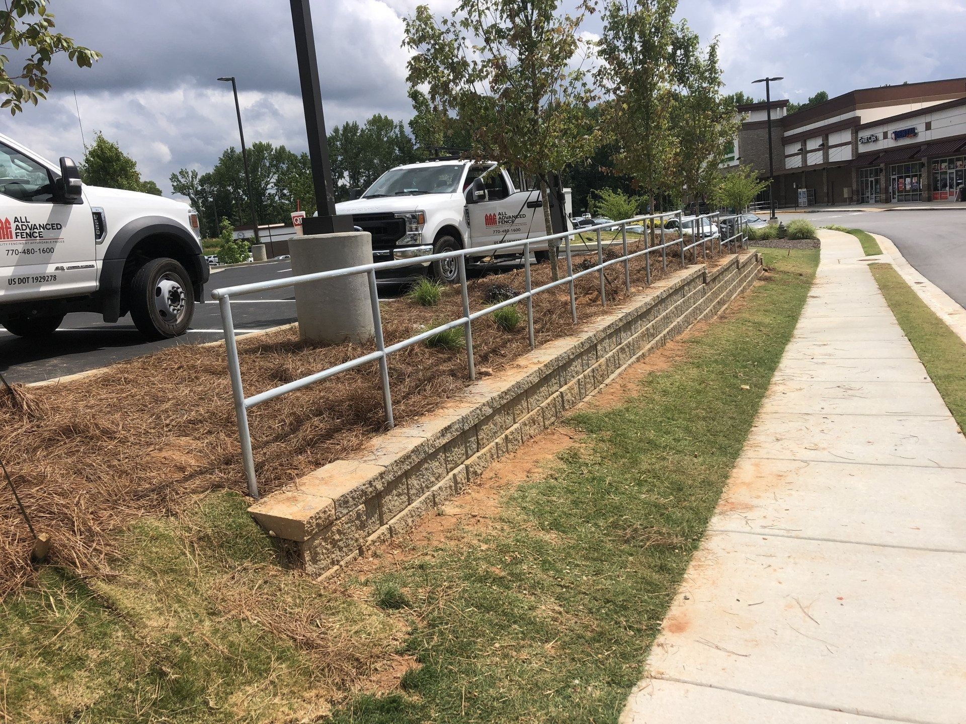 A landscaped area with retaining walls, a metal railing, and a sidewalk next to a parking lot with work trucks.