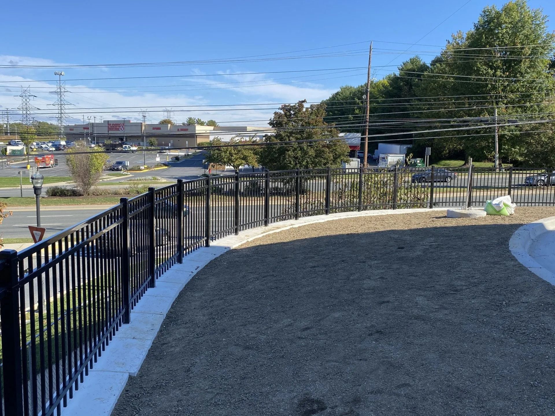 Black metal fence bordering a gravel area with a view of a parking lot and trees under a blue sky.