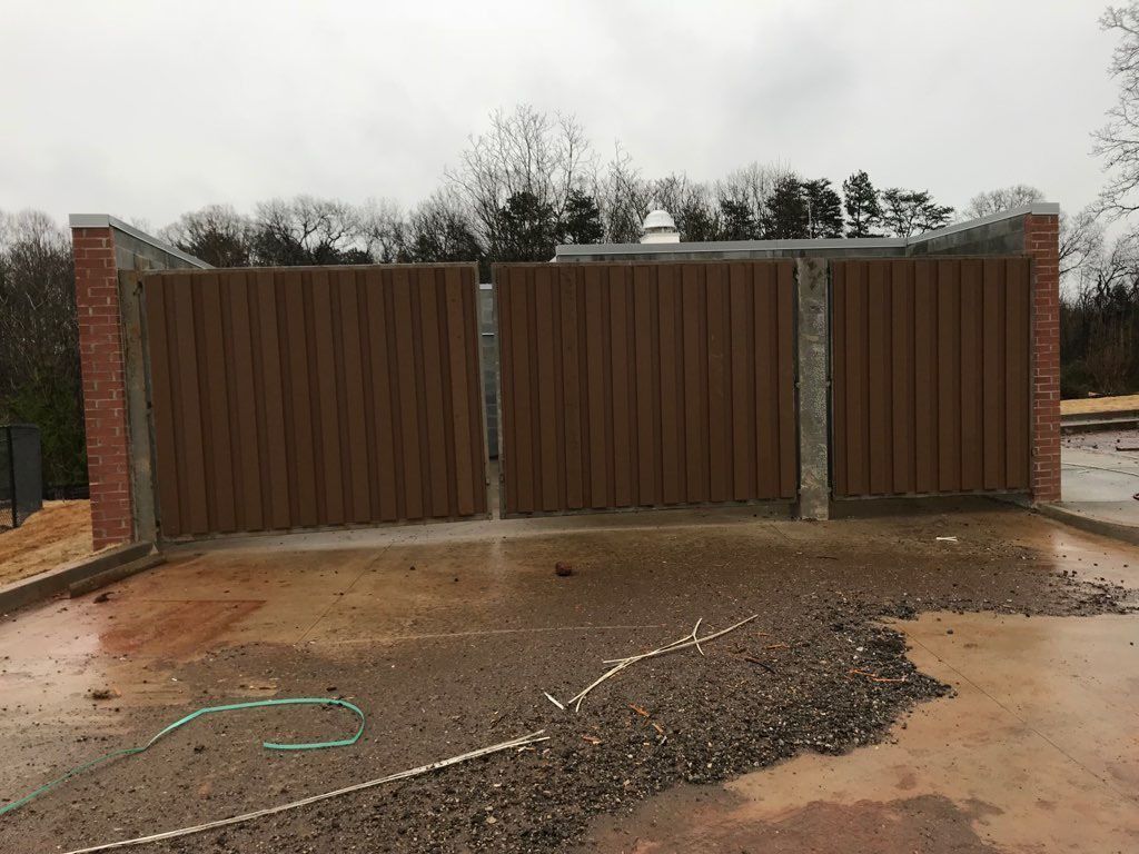 Brown double gate with vertical slats, brick and concrete posts, gravel, overcast day.