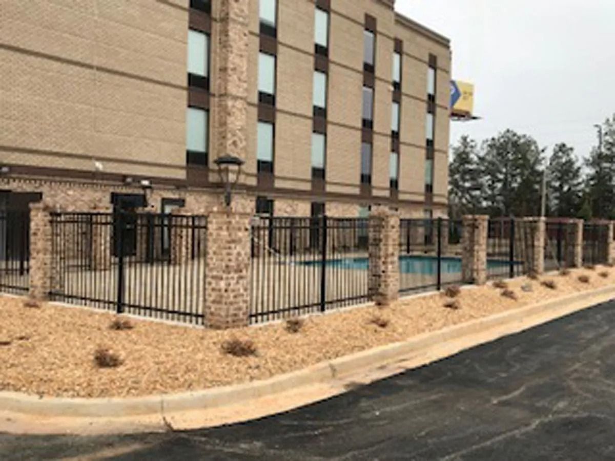 Hotel with a black fence, brick columns, and a pool. Building is light brown, set on tan gravel.