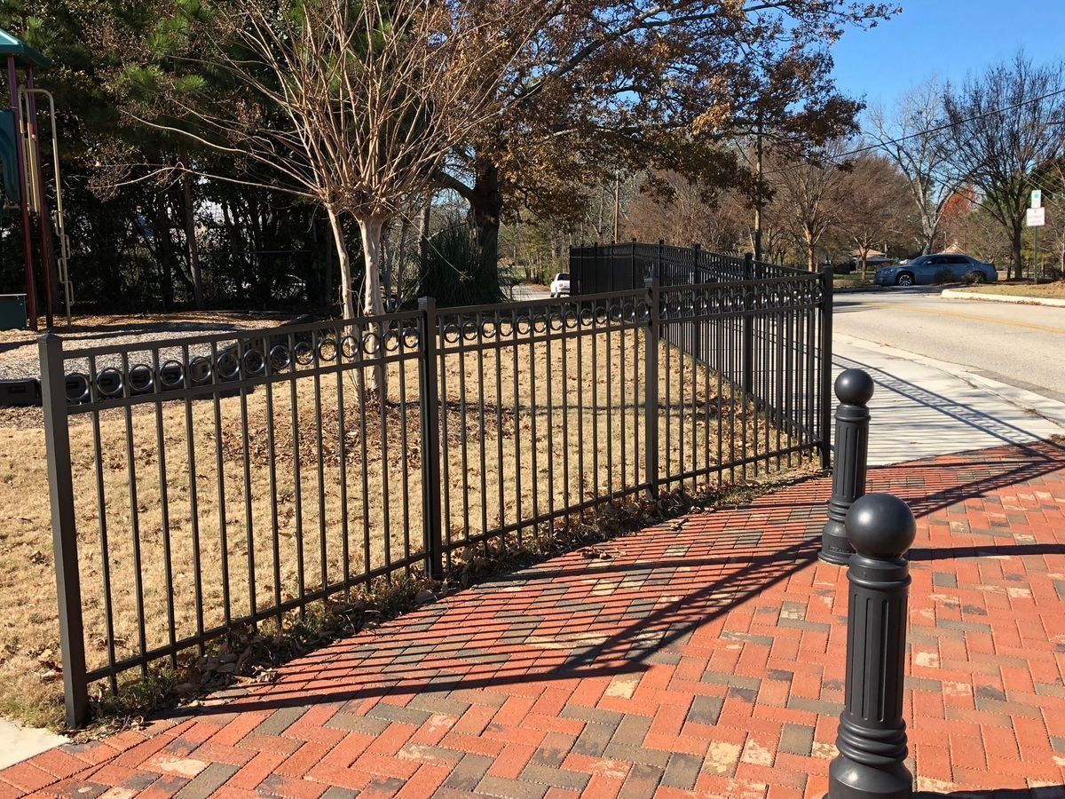 Black metal fence bordering a brick sidewalk and road; trees in the background.