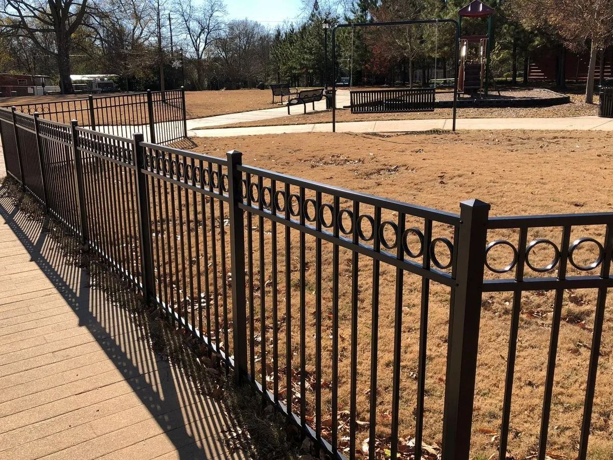Black metal fence surrounds a playground with brown ground and trees.