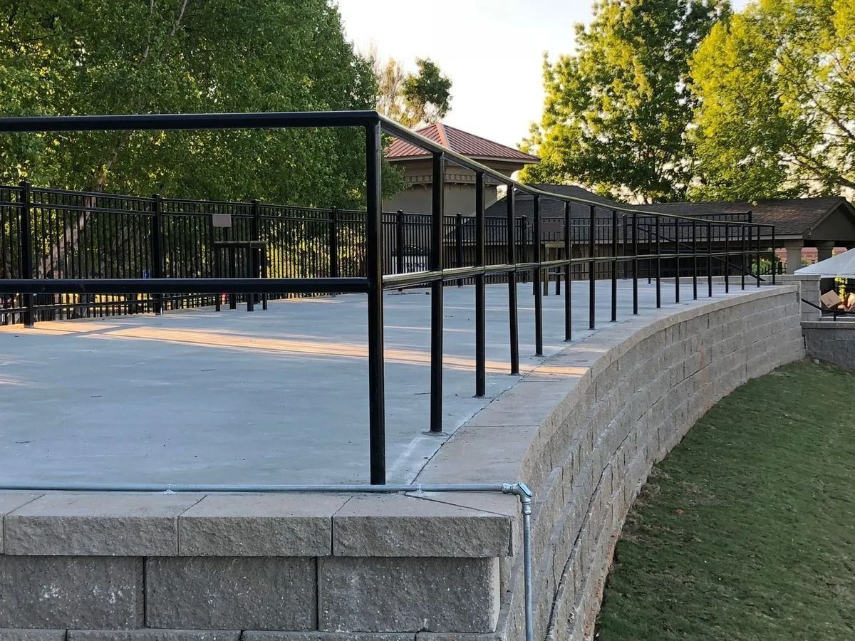 Concrete patio with black metal railing atop a curved, gray block retaining wall. Green grass and trees in background.