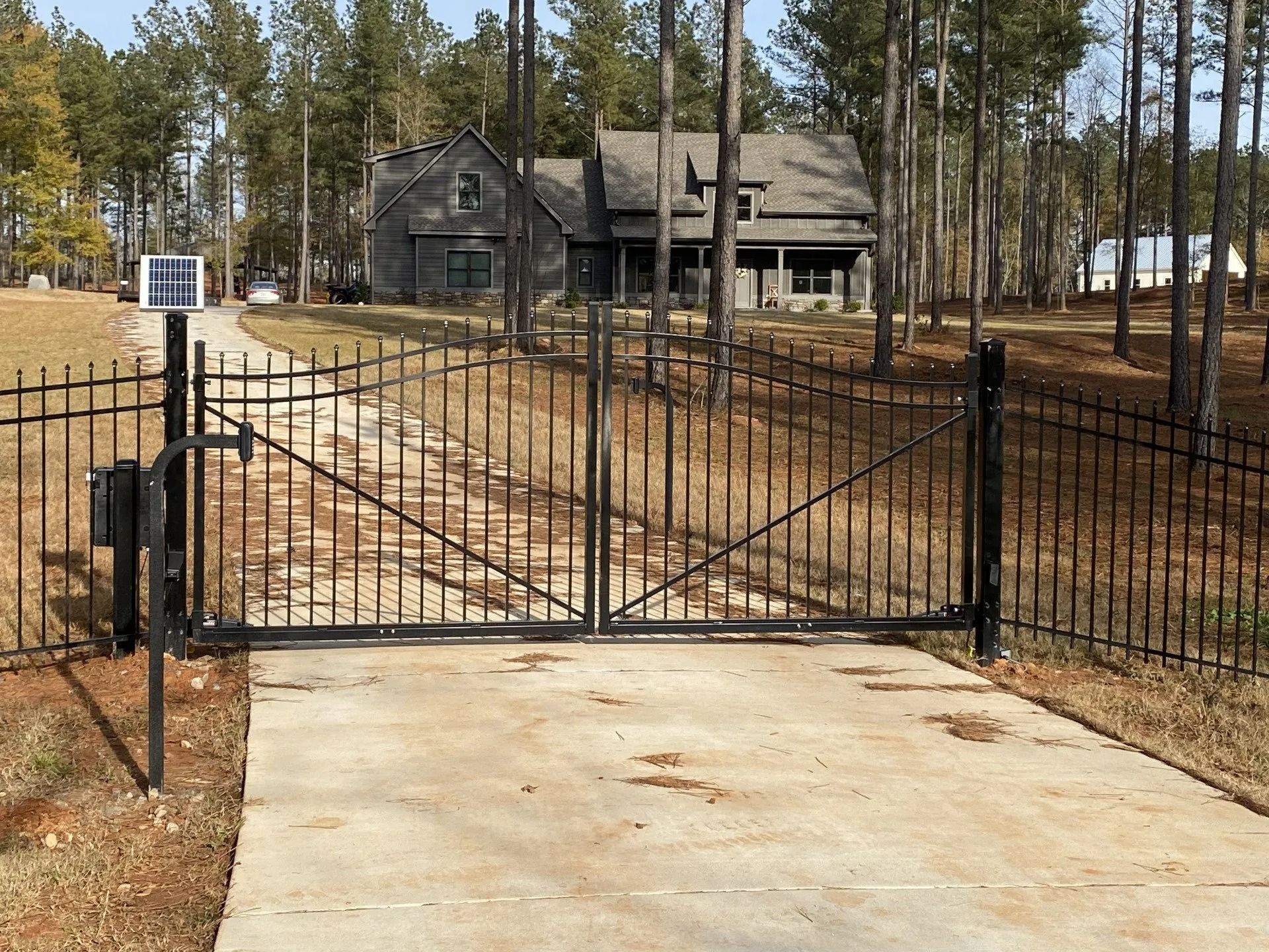Black metal gate on a concrete driveway leading to a gray house in a wooded area.