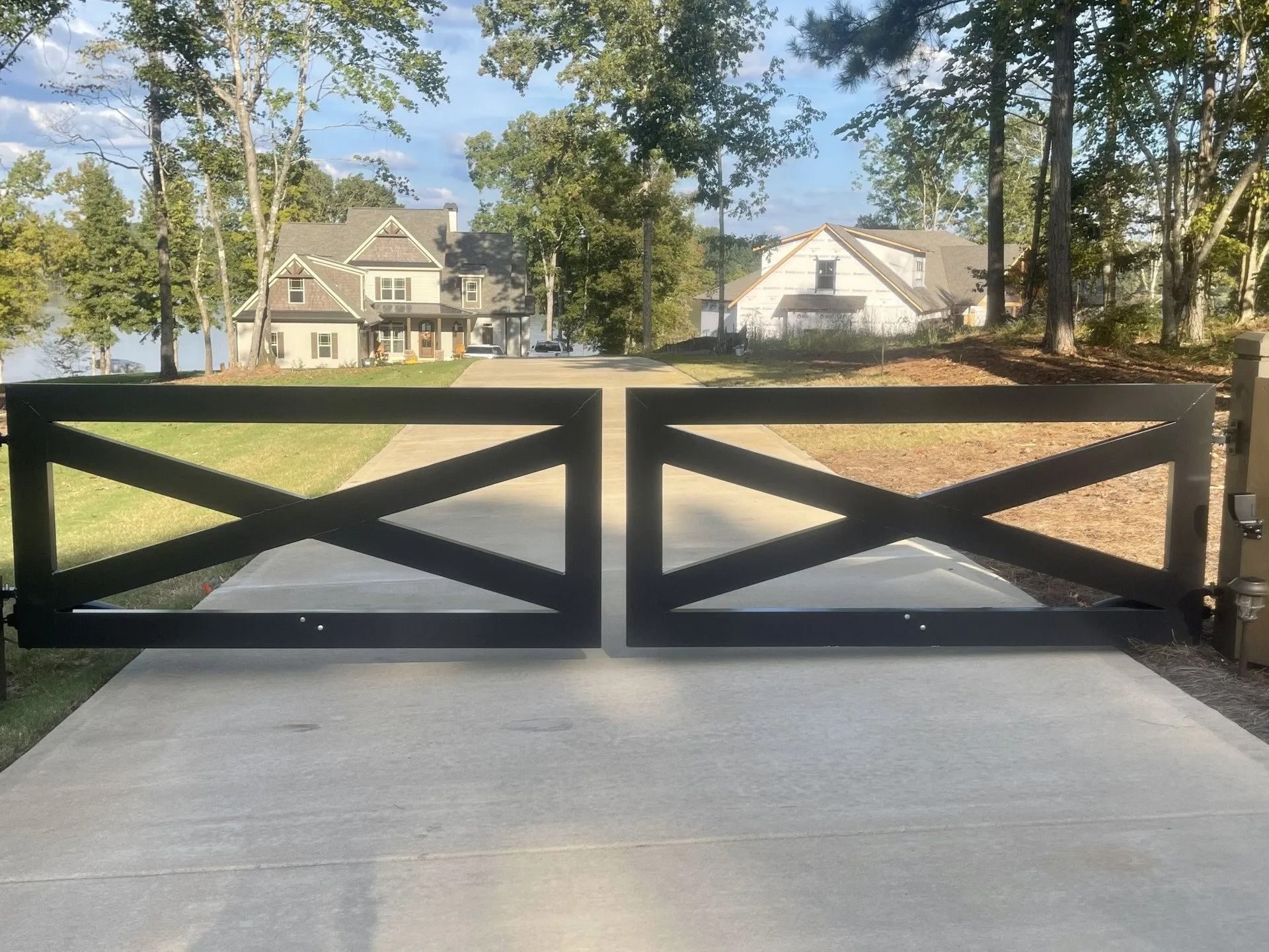 Black X-style driveway gates open on a concrete drive leading to houses near a lake.