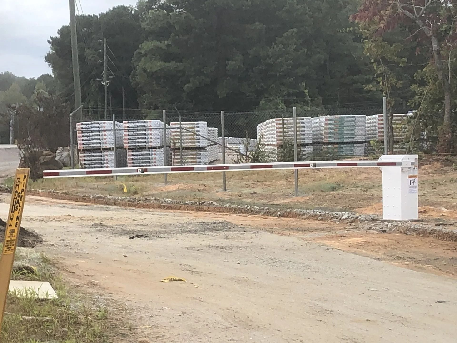 A dirt road with a gate, stacks of construction materials, and a forested backdrop.