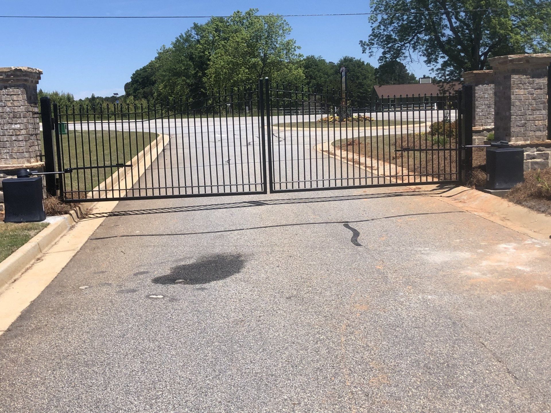 Black metal gates on a paved road, flanked by stone pillars, leading to a green area under a blue sky.