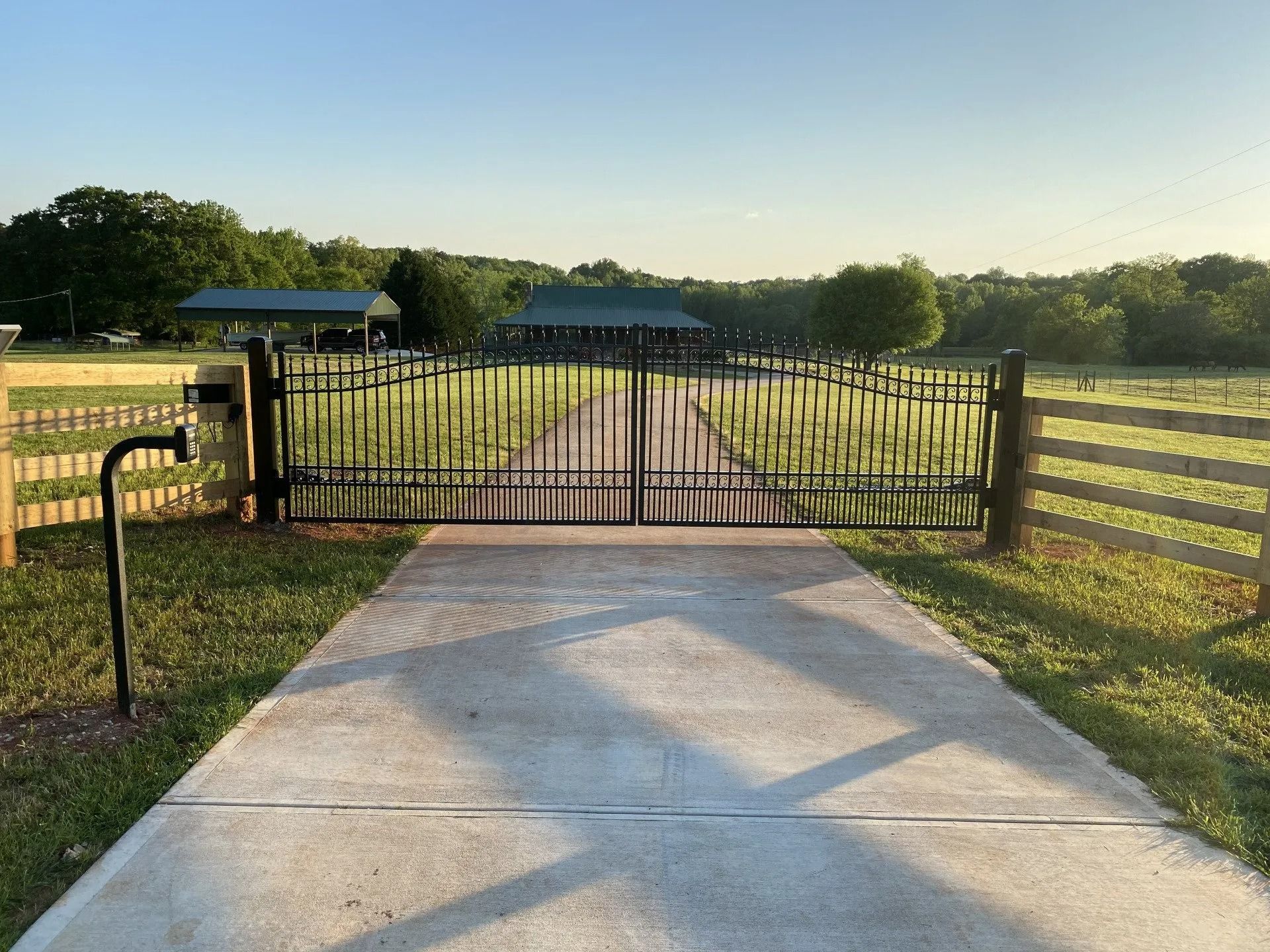 Black gate on a concrete driveway, leading to a home; wooden fence on either side.