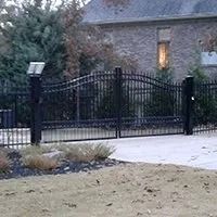 Black metal driveway gate with brick house and trees in the background.