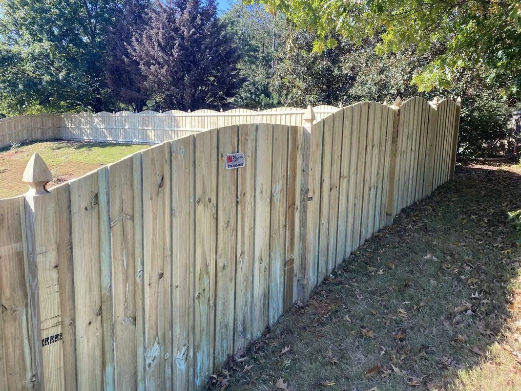 Wooden fence curving along a yard, with arched top and decorative finials, against trees and blue sky.