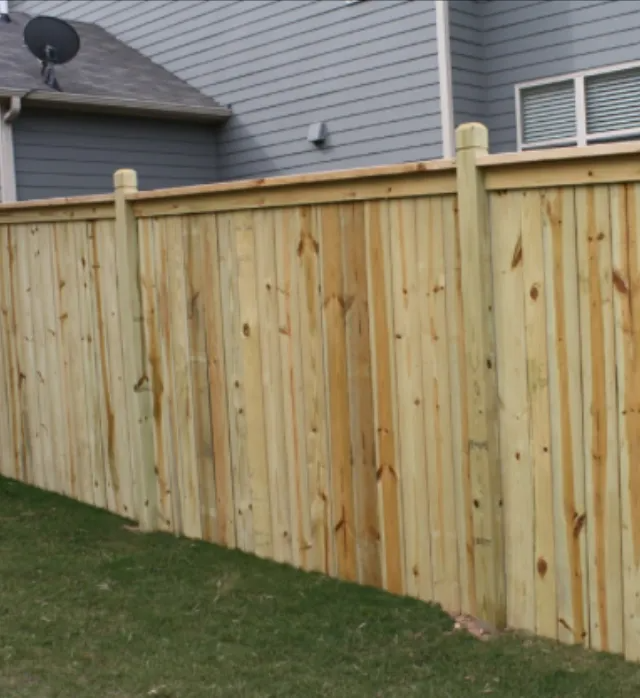 Wooden fence in backyard, green grass in front, gray siding of house behind.