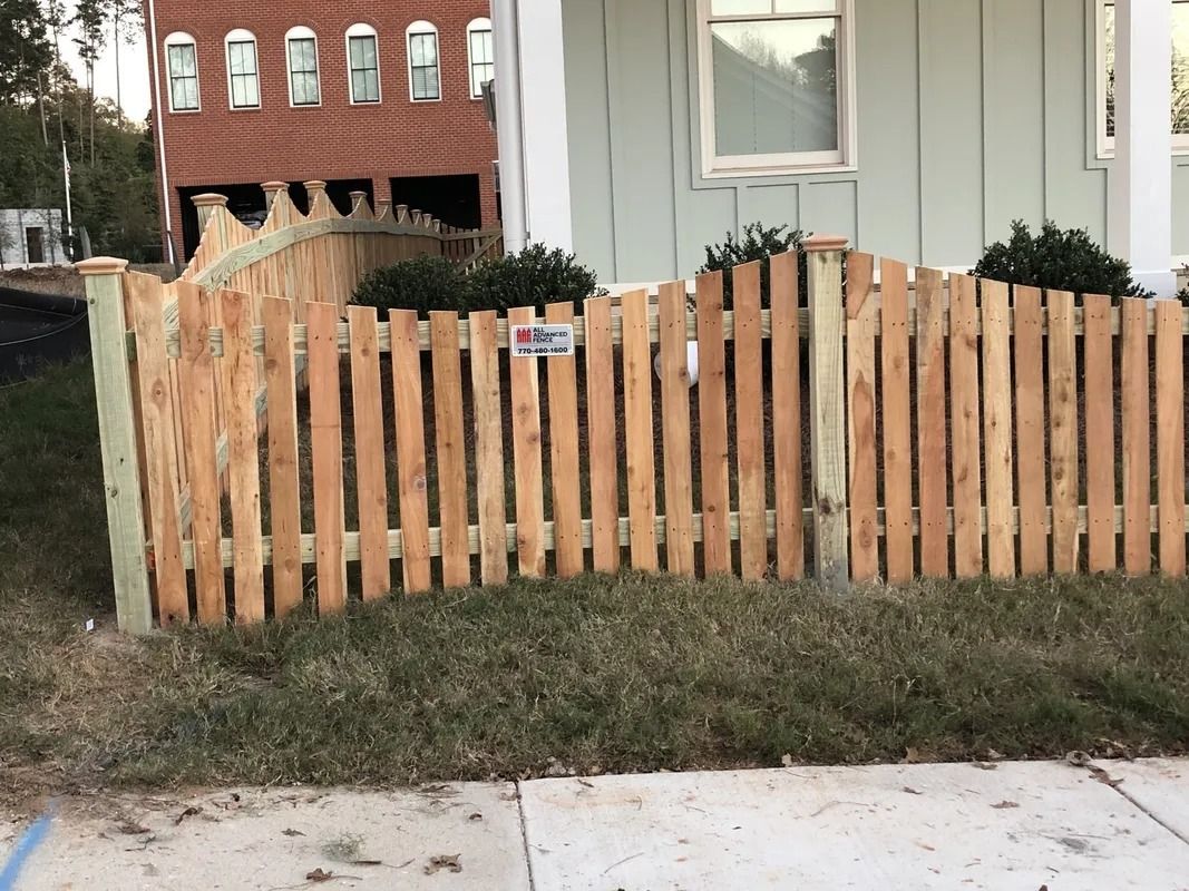 Wooden picket fence in front of a light green house, with a brick building visible in the background.