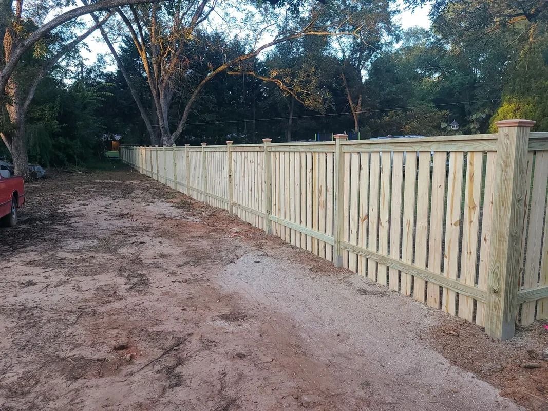 Wooden fence along a gravel path in a yard, trees in the background.