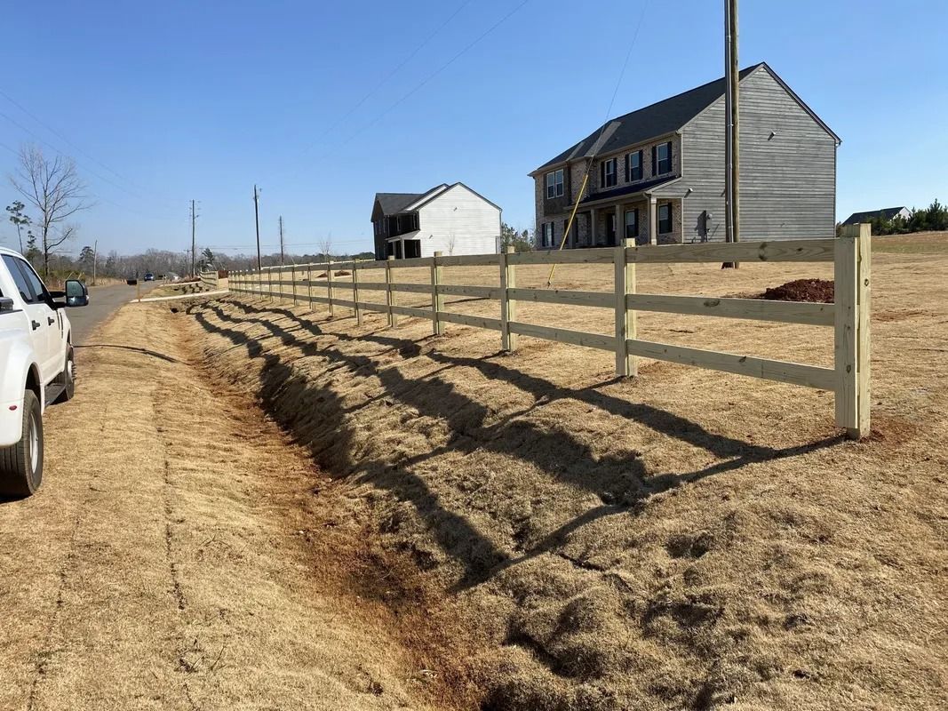 A white three-rail fence alongside a ditch in a field with houses under construction in the background.