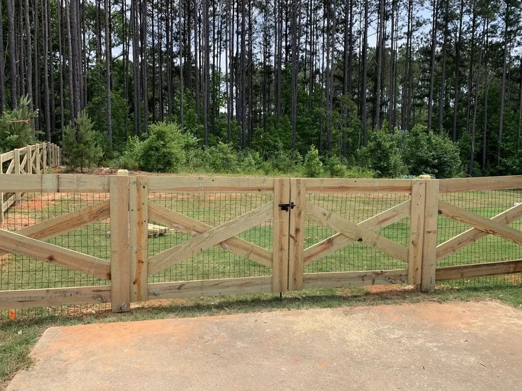 Wooden fence with gate, featuring X-shaped bracing and wire mesh. Green foliage and trees in background.