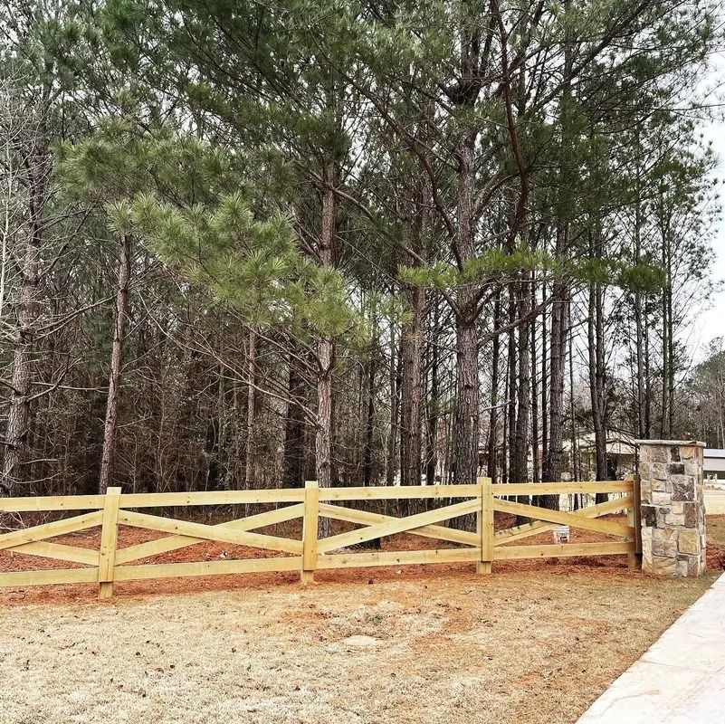 Wooden cross-buck fence with stone pillars in front of a forest of trees.