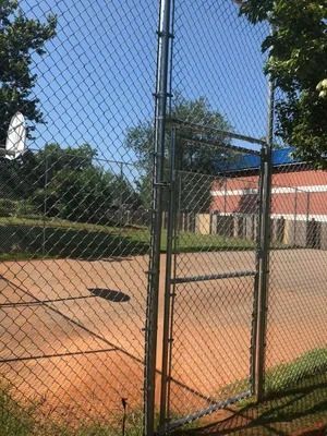 Chain-link fence with a gate, enclosing a basketball court and a red brick building on a sunny day.
