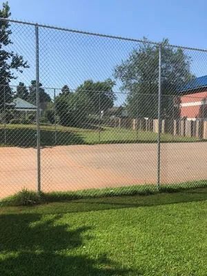 Chain-link fence in front of a dirt field and some trees, under a bright, sunny sky.