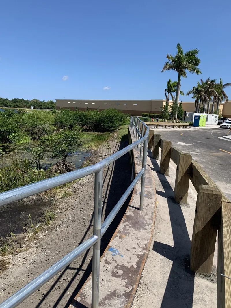 Concrete walkway with metal railing next to a body of water and a building on a sunny day.
