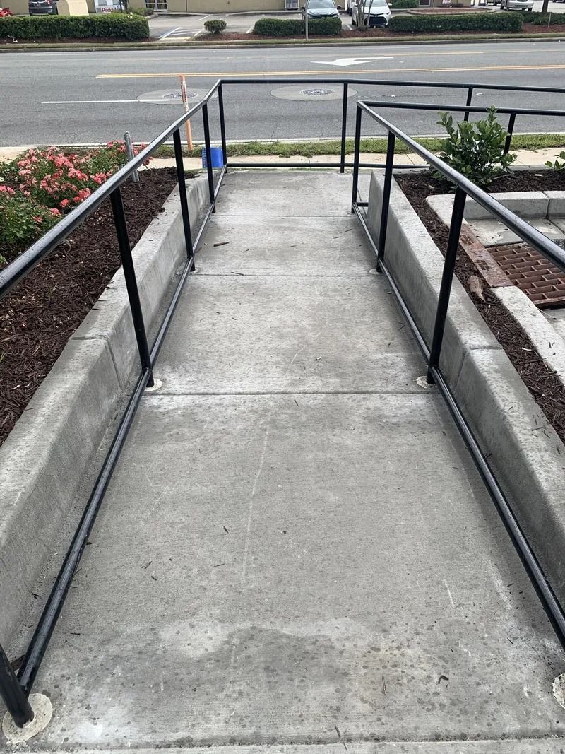Concrete ramp with black handrails, bordered by flowerbeds, leading to a street.