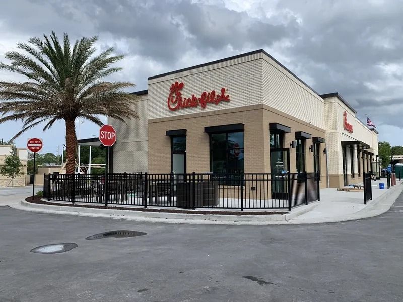 Chick-fil-A restaurant exterior with a palm tree, sidewalk seating area, and overcast sky.