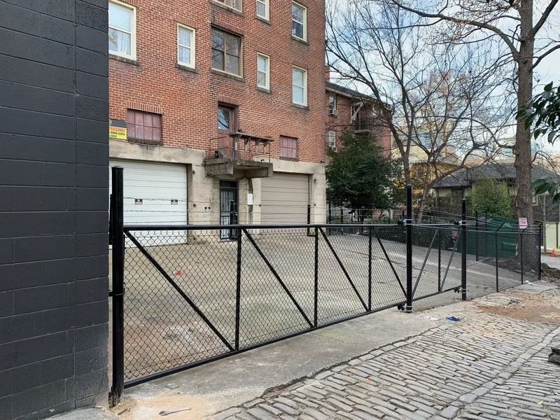 Black metal fence encloses a parking area next to a brick building with garage doors. Paved street.