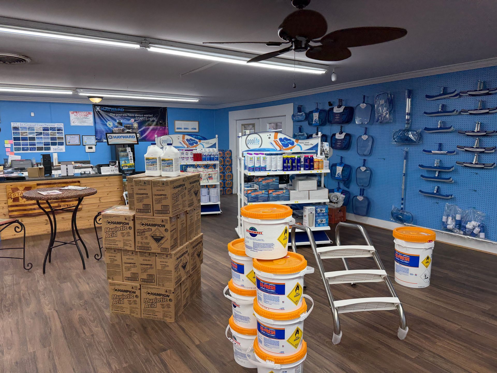 Pool supply store interior with products on shelves and floor, including chemicals, equipment, and a ladder.