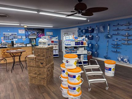 Pool supply store interior with products on shelves and floor, including chemicals, equipment, and a ladder.