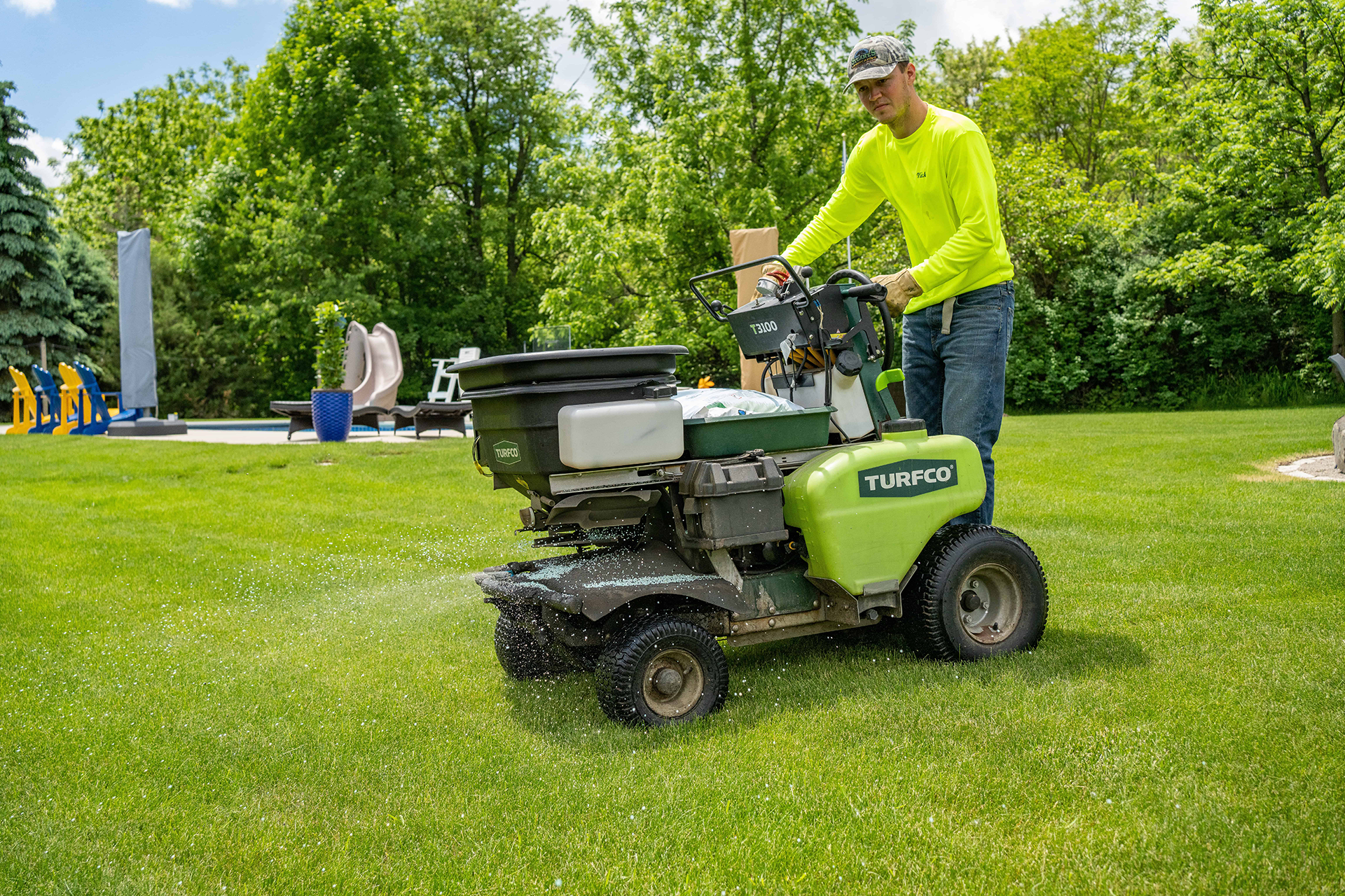 Man operating a green lawn spreader, spraying grass on a sunny day.