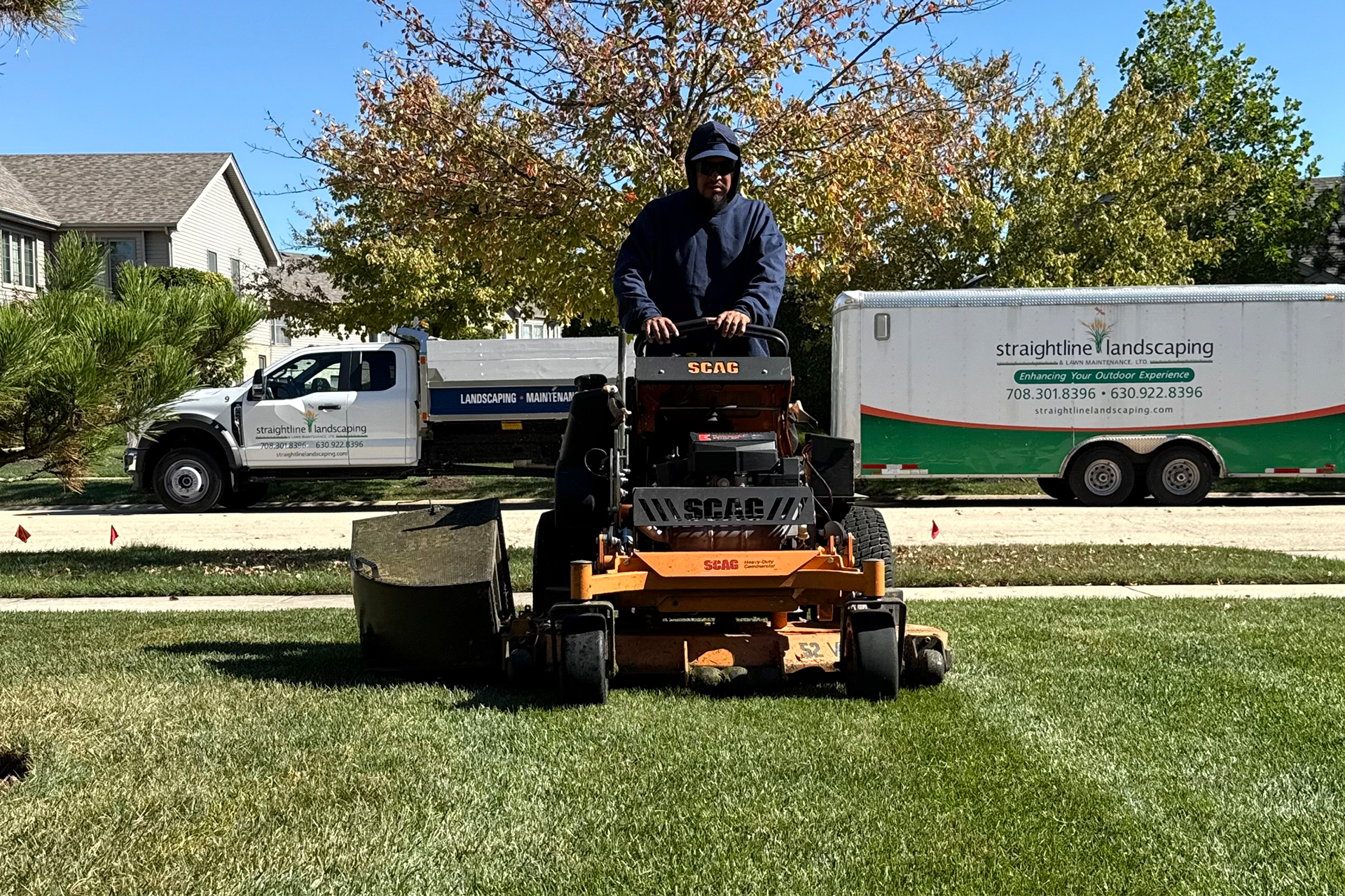 Person operating a commercial lawn mower, cutting grass in a residential yard. A truck and trailer are visible in the background.