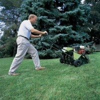 Man pushing an aerator across a green lawn, near trees.