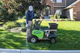 Man using a green fertilizer spreader on a lawn near a house.