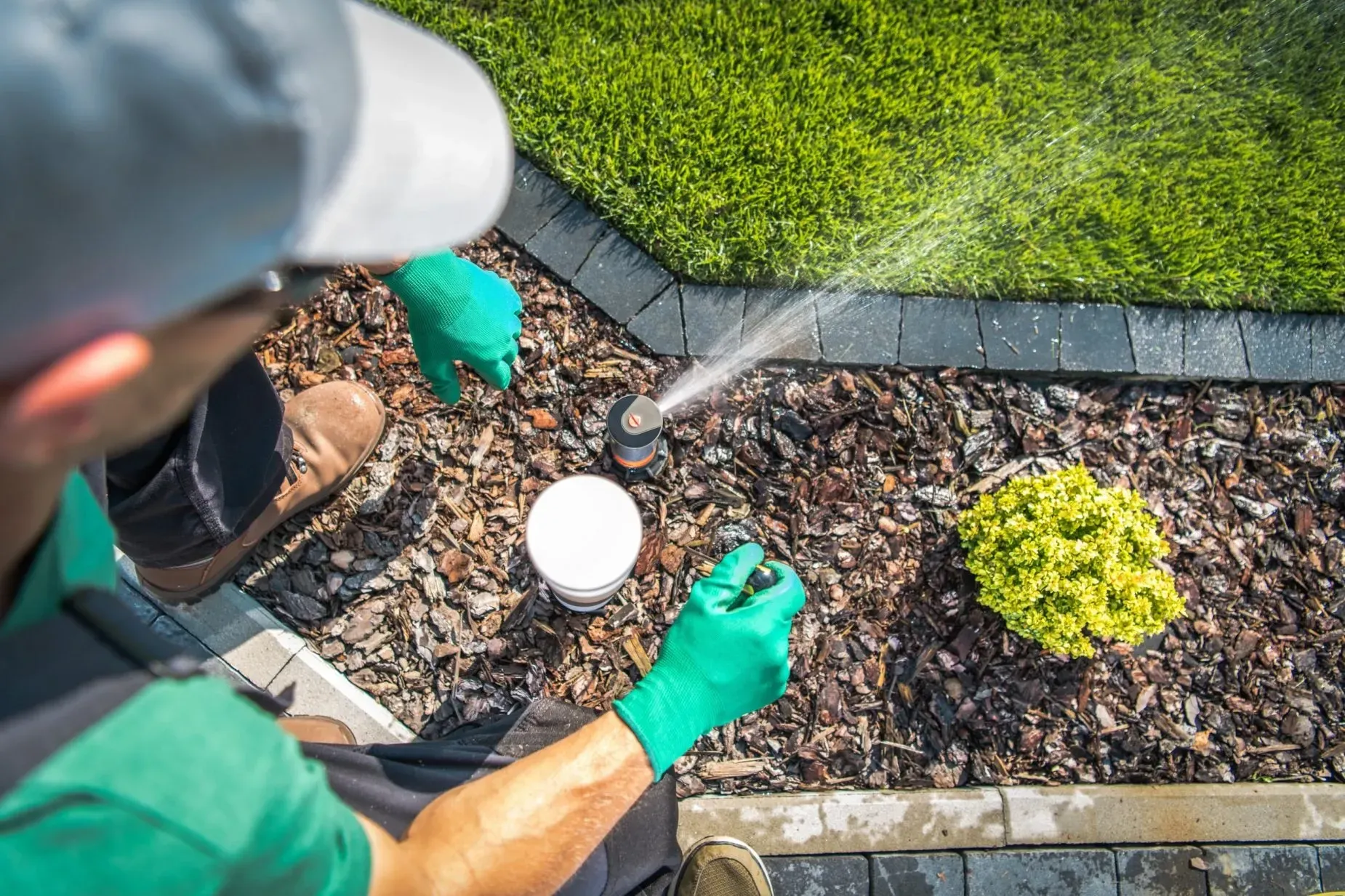 Person in green gloves adjusting a sprinkler in a mulched garden bed.