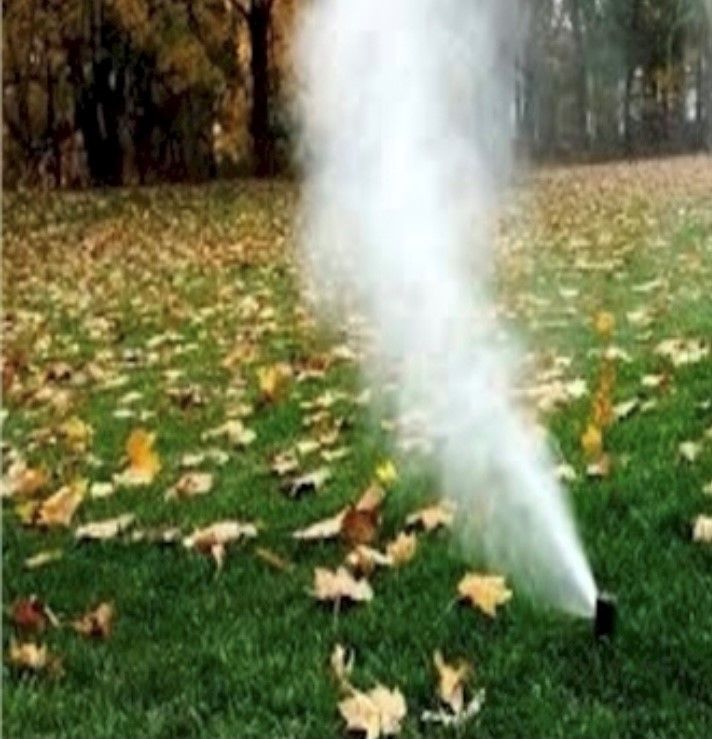 A sprinkler sprays water upwards, creating a white cloud over a green lawn covered in fallen leaves.