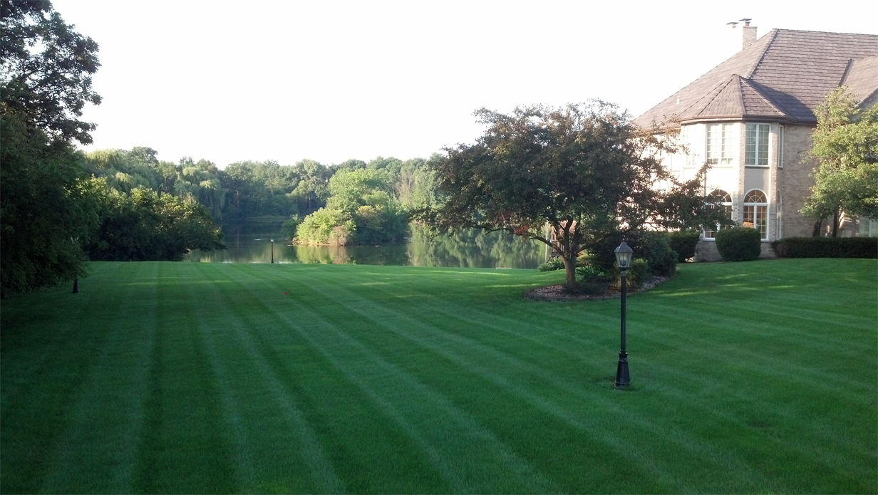 Lush green lawn with striped mowing pattern, leading to a lake. A large house sits to the right, trees in the background.